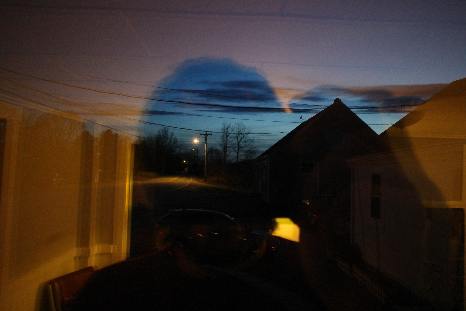 Collage-esque photograph that looks like a double exposure with Alex Silver's silhouette, but is the mixture of a lamp-lit window reflection and the quiet, empty street of a very early morning, mostly blue, yellow, and black colors, by Maine musician Alex Silver.