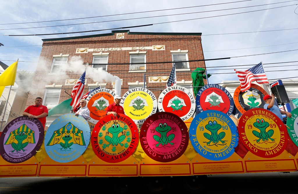 "The La Kermesse float passes on Jefferson Street in Biddeford as riders wave to spectators during a parade kicking off the 36th annual La Kermesse Franco-Americaine Festival. — Staff photo by Derek Davis." This is a quotation from the Portland Press Herald website describing the original photo. The float includes many of the circular emblems for the festival, which include a green frog and the year, as well as "C'est le temps," the motto of La Kermesse.