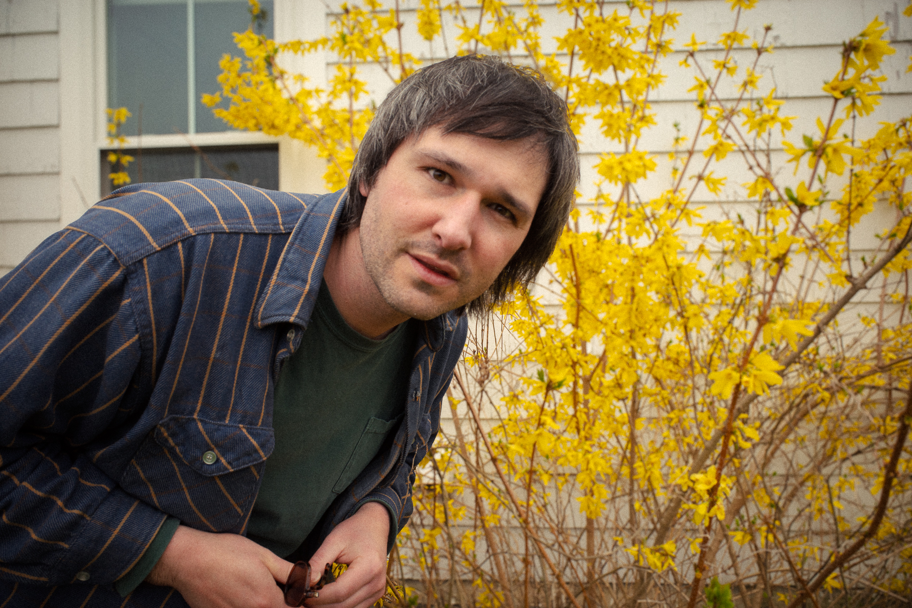 Alex Silver staring at the camera in front of a blooming yellow bush and a house with white siding.
