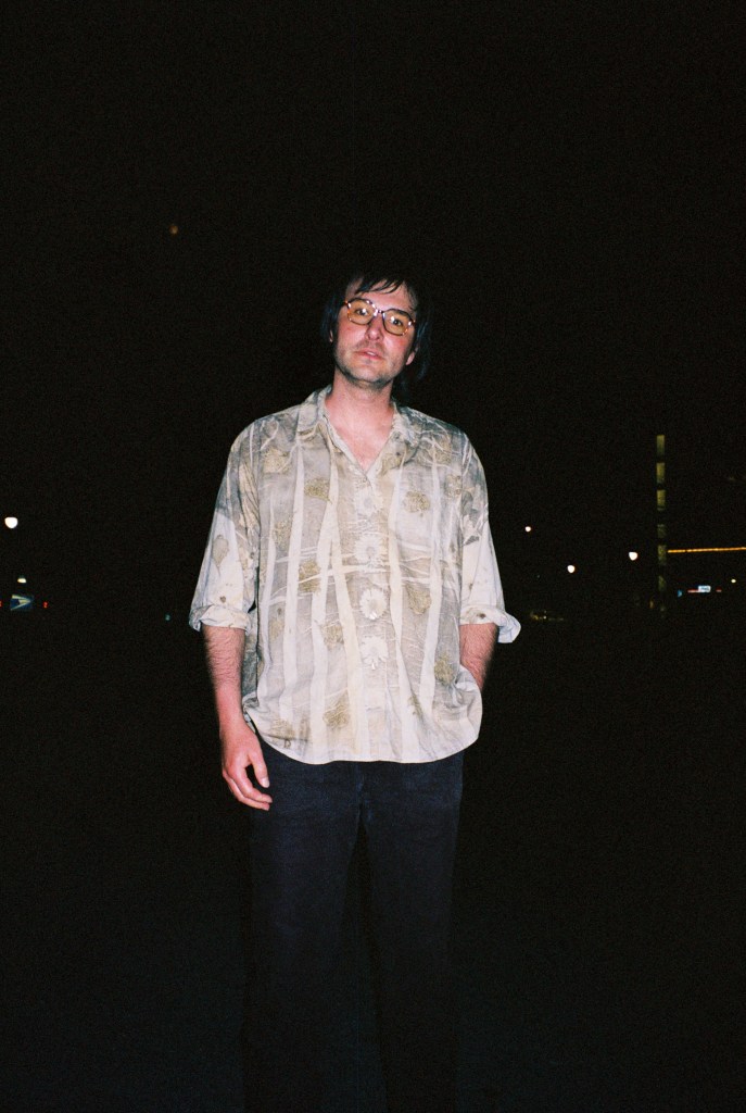 Alex Silver outside the Apohadion Theater, taken on a film camera with flash at night. Silver wears an eco-dyed shirt with open collar, patterned with vivid forms of flora, and glasses with transition lenses slightly yellow-tinted, with one hand in a pocket and the other out.