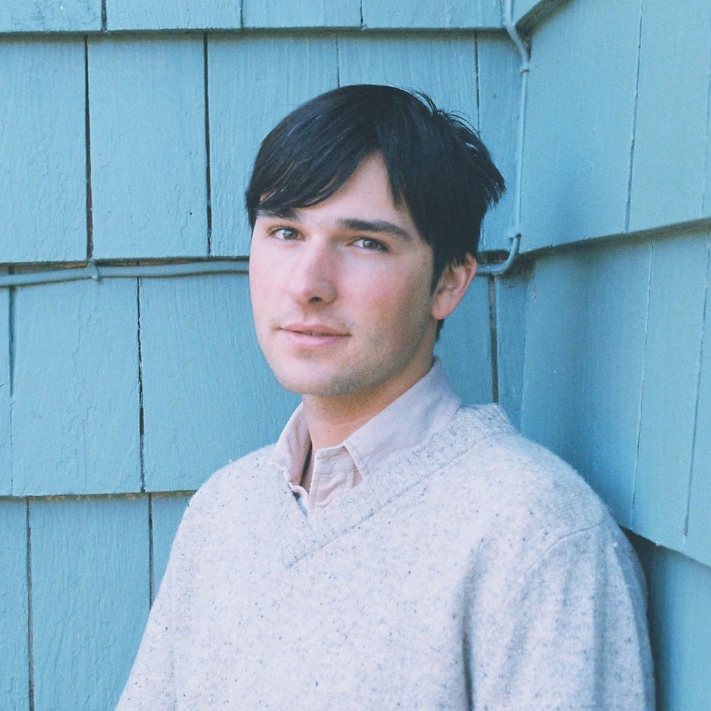 Alex Silver looks directly at the camera, standing at a slight angle in front of teal siding. He wears a cream-colored sweater over a vintage collared shirt.