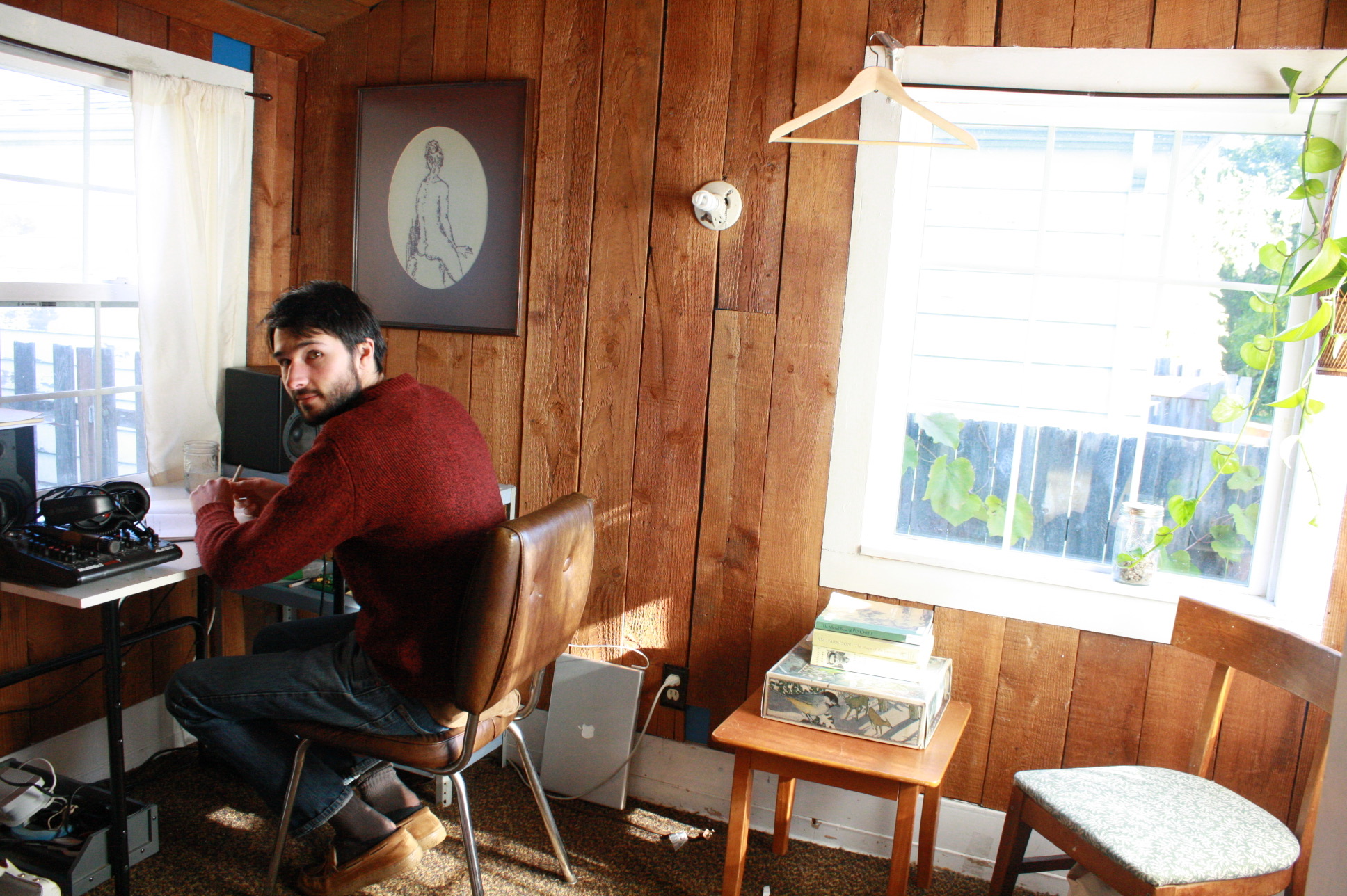 Alex Silver looking at the camera from his writing desk in a small room in Olympia with wooden plank walls. On the desk is recording equipment and a notebook. Light is streaming through the windows.