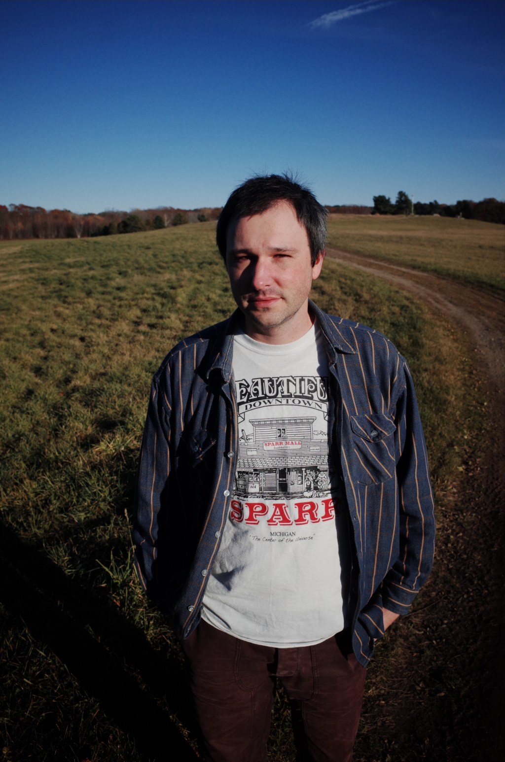 Alex Silver in a white graphic shirt with black and red text, covered in a blue overshirt with gold stripes, standing in a field with a dirt road passing into the background.